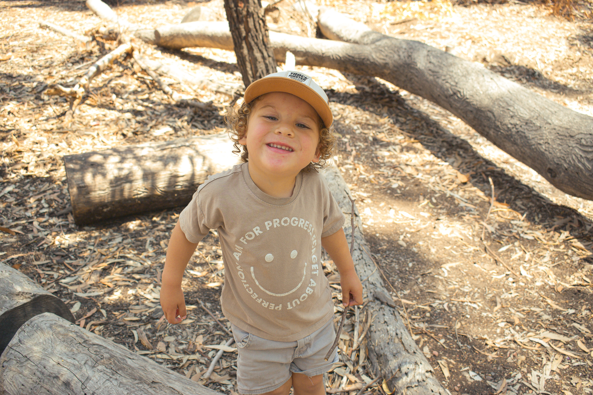 Smiling child with curly hair and a cap standing among fallen logs in the forest