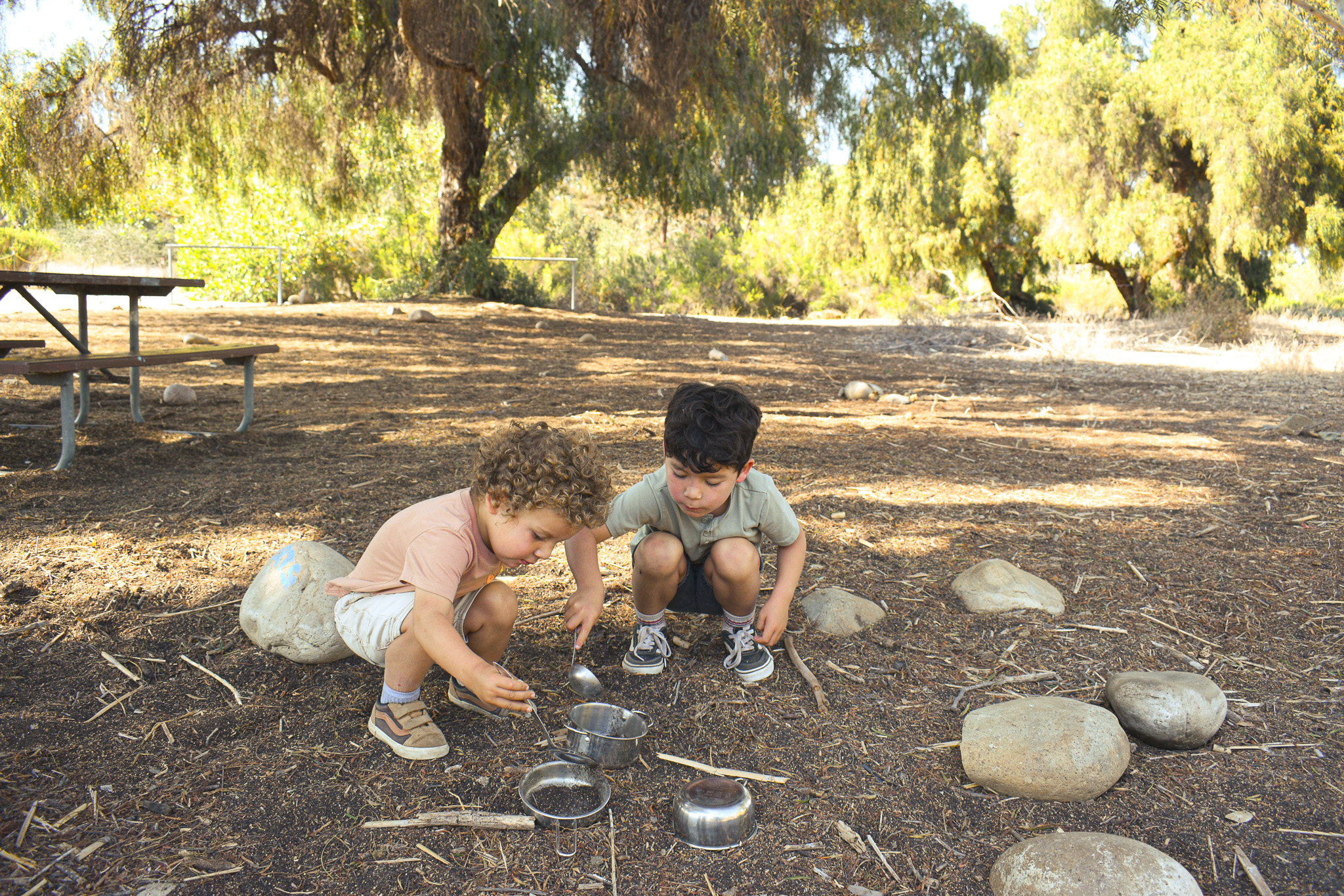 Two children crouched together playing with pots and sticks in the dirt under shady trees